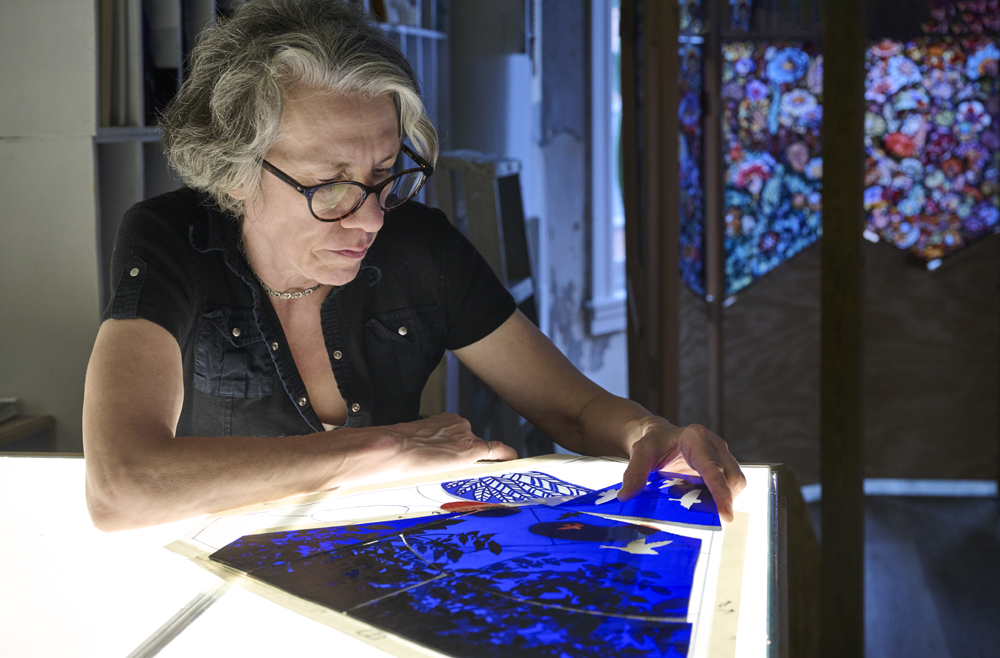 Artist Judith Schaechter sits at a table piecing together sections of glass.